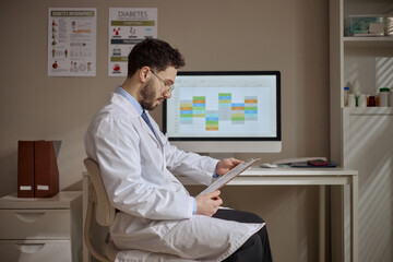 Fototapeta premium Young adult Middle Eastern male doctor reviewing clipboard at computer with schedule on screen in clinic, managing patient records. Useful for healthcare administration, medical workflow, telehealth