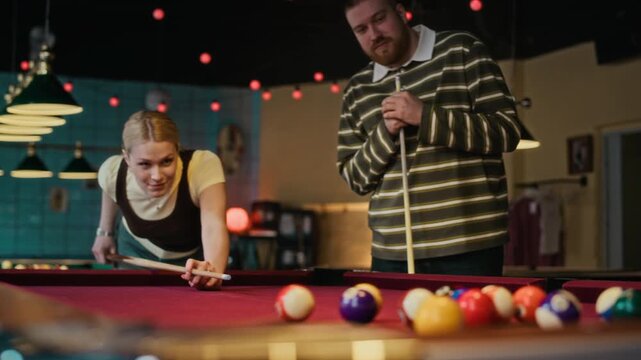 Rack focus shot of young adult woman hitting opening shot aiming at racked balls on red felt table while gathering with friends at pool hall