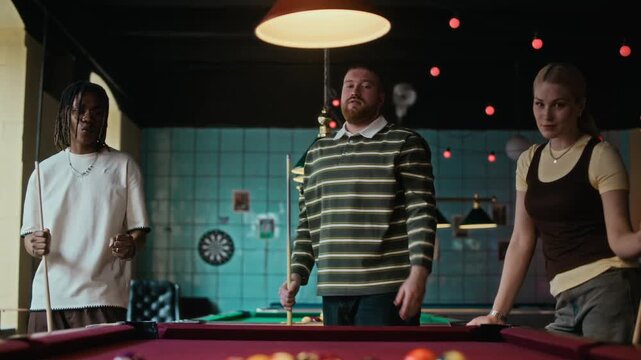 Tilt down shot of young adult man striking break shot in billiards game while hanging out with friends at pool hall