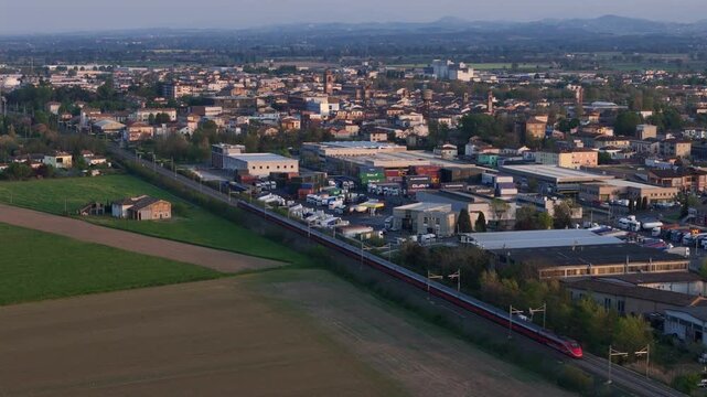 Trenitalia Frecciarossa high speed train on Milano Bologna AV line passing Fiorenzuola d&rsquo;Arda Piacenza Emilia Romagna Italy with town skyline and distant Ligurian Apennines ridge, drone tracking ascen