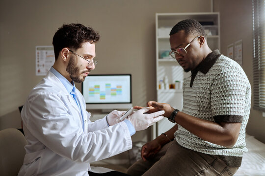Young adult Black man receiving finger prick testing for blood glucose monitoring from Caucasian male doctor during clinic visit. Useful for healthcare, diabetes screening, medical consultation