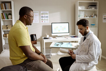 Fototapeta premium African American young adult man consulting male doctor, discussing symptoms during clinic appointment. Useful for healthcare services, patient care, medical consultation marketing