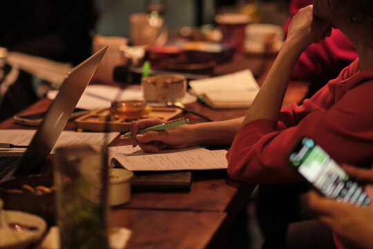 Close side view of a participant writing notes with a green pen at a warm workshop table, with laptop and drinks nearby. Adult learning and group discussion support focus, skills, and personal growth.