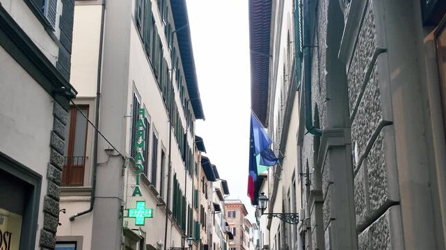 Charming narrow street scene in Italy with traditional architecture, pharmacy neon sign, Italian and EU flags, soft daylight, and textured building facades in a steady wide shot.