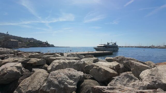 Tranquil harbor scene with large rocks in foreground, a luxury yacht docked by the pier, sunlit water, blue sky, and distant hillside. Wide angle, steady daylight footage. (198 characters)
