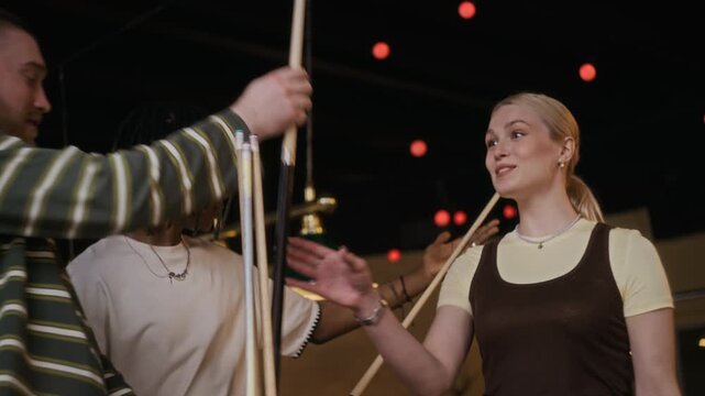 Tilt up medium shot of cheerful young adult men and woman taking cues before playing billiards game while getting together at pool hall