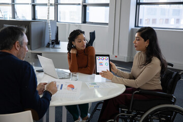 Diverse team reviewing reports at table in office, wheelchair woman presenting tablet, copy space