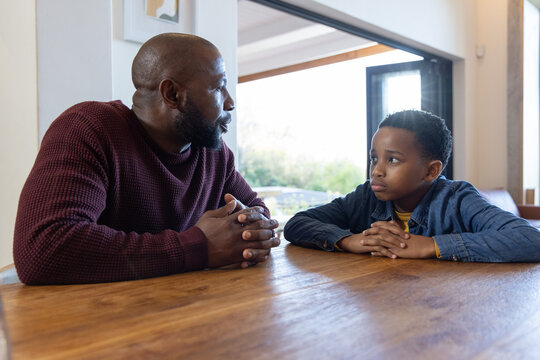 African American father and son sitting at wooden table near sliding door, maroon knit having talk