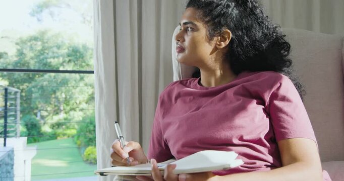 Adult woman journaling at home with pen in notebook, sunlight drawing gaze toward garden window