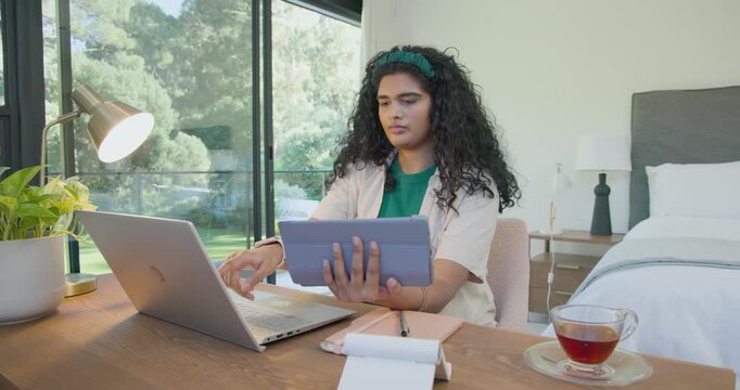 Indian woman at desk referencing tablet, placing on stand, typing on laptop for work, sipping tea