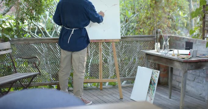 Senior Asian man wearing smock apron holding brush starting piece with light strokes on easel porch