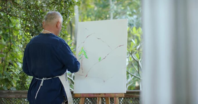 Senior Asian man in apron painting at easel on porch, moving sketch into green color