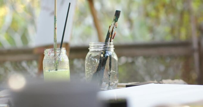 Camera is sliding and pulling focus, revealing two glass jars of brushes on table, showing canvas