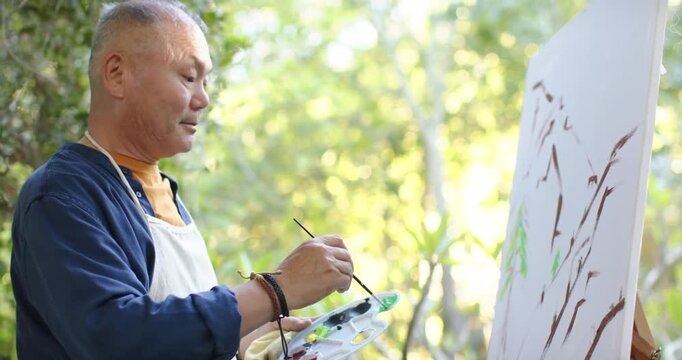 Senior man at easel wearing apron mixing on palette and painting leaves for nature scene outdoors