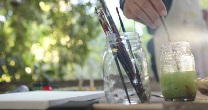 Senior man entering frame stirring brushes in green jar by canvas on outdoor table prepping artwork