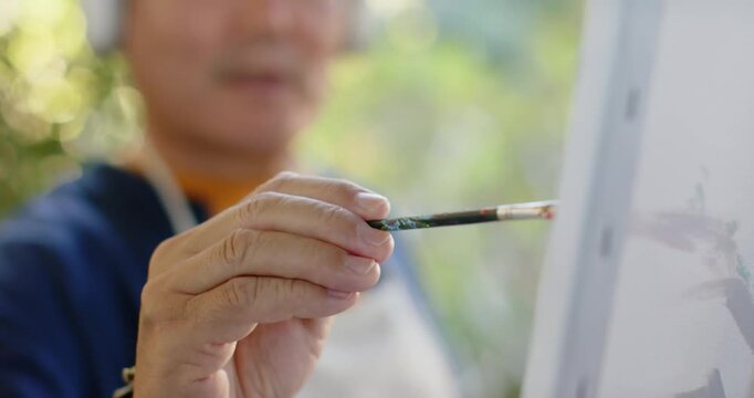 Senior Asian man painting canvas on easel in garden, making marks showing leaves with headphones