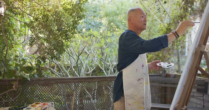 Senior Asian painter on porch painting at easel in stained apron studying dappled light, copy space