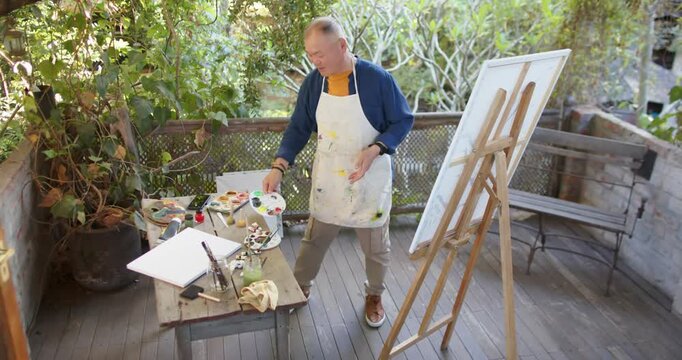 Senior Asian man in apron, surveying area, preparing palette, checking phone for reference on porch