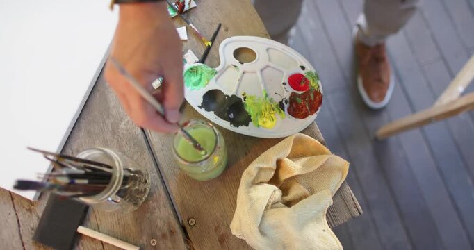Reaching for palette Senior Asian man rinsing brush in jar, wringing rag on wooden table preparing
