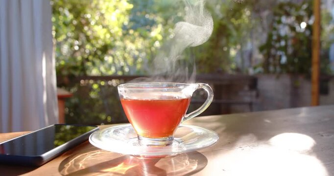Glass teacup is releasing steam as hot red tea is cooling on wooden table, phone nearby