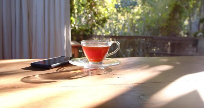 Glass teacup sitting on glass saucer on wooden table while sunlight is shifting due to breeze