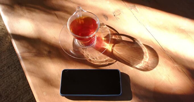 Sunlight sliding across sunlit wooden table is casting ring from teacup and nudging phone clockwise