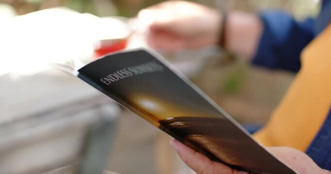 Right hand is initiating fanning ENDLESS SUNSETS booklet pages to reveal title on porch near cup