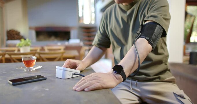 Senior Asian man pressing BP monitor, tightening cuff on left arm at dining table, checking health