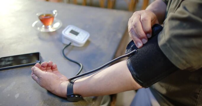 Senior Asian man sitting at table preparing blood pressure reading, placing cuff checking monitor