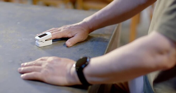 Senior Asian male tapping then clipping white fingertip sensor to finger at table, checking oxygen