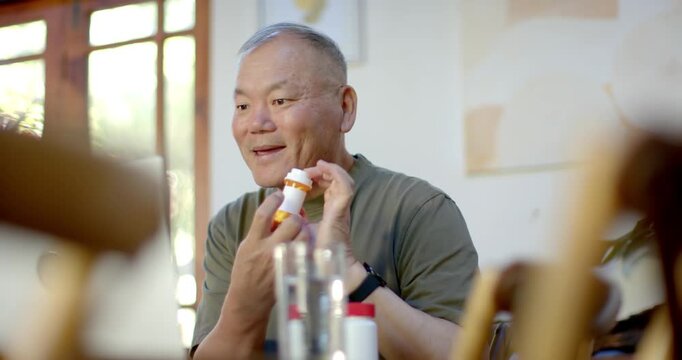 Senior Asian man prompted by offscreen cue reaching and inspecting orange pill bottle at home table