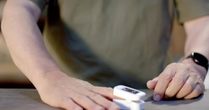Senior Asian male bringing white oximeter to left finger, clipping and checking oxygen on table
