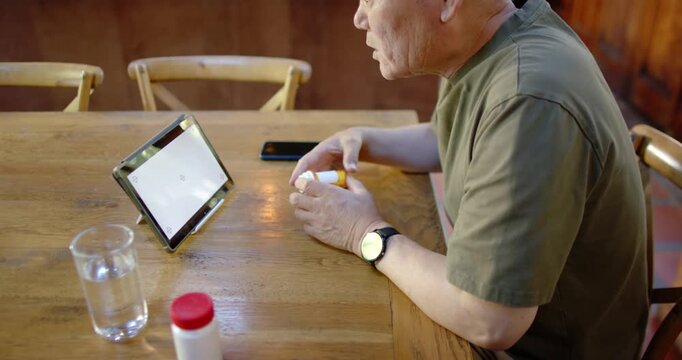 Senior Asian man sitting at dining table consulting tablet on stand checking pill bottle for dosage