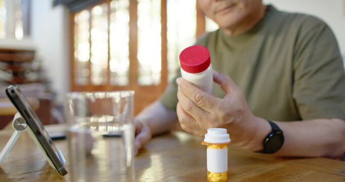 Senior Asian male checking tablet at table, grabbing redcap bottle, reading label confirming meds