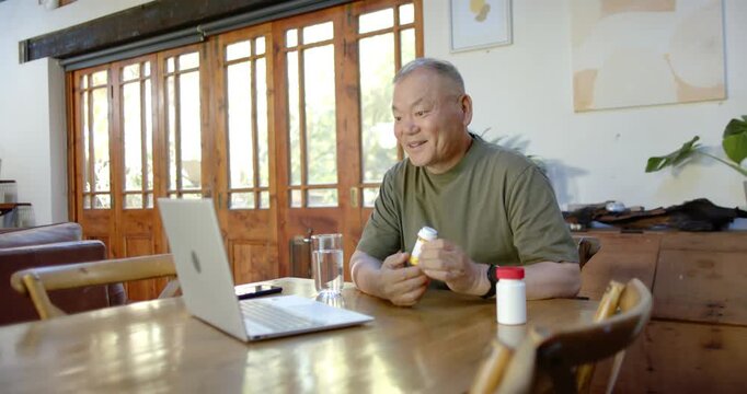 Senior Asian man at table lifting yellow Rx bottle toward laptop on telehealth consult, copy space