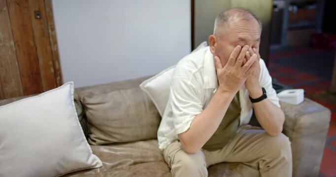 Senior Asian man sitting on couch feeling tired covering face yawning clasping chin glancing right