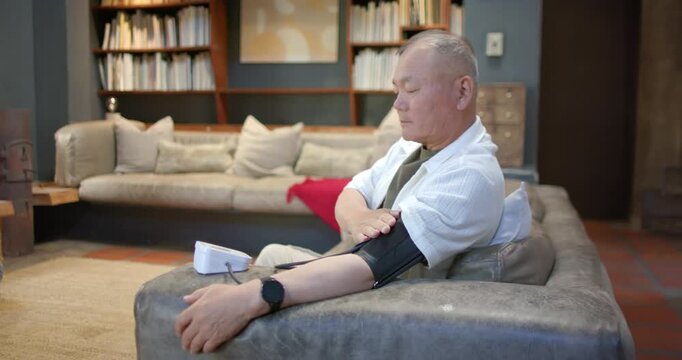 Senior man seeing cuff, rolling sleeve and starting monitor for BP reading on sofa, copy space