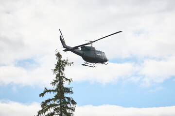 Fototapeta premium Military Helicopter Flying Over Pine Tree Against Partly Cloudy Blue Sky On Patrol