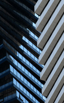A tight, close-up abstract of two modern building facades meeting in a geometric composition. The shot focuses on the rhythmic contrast between the dark blue glass window grid and the sharp