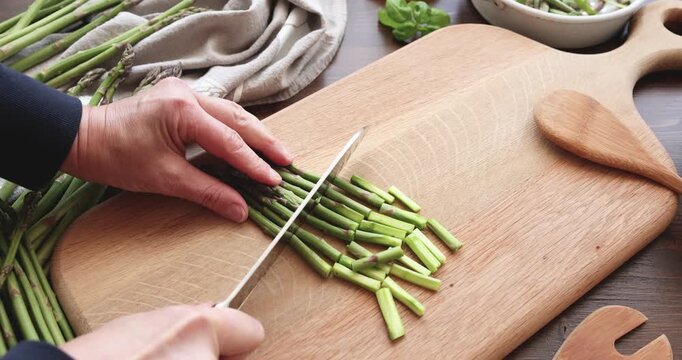 Woman hands cutting green asparagus with sharp knife on wooden board. Chef preparing fresh organic vegetables for healthy meal in kitchen. Culinary process of slicing raw ingredients for delicious