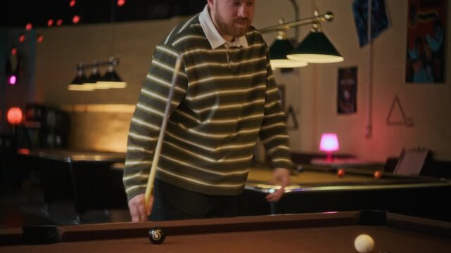 Tracking shot of focused young adult man walking beside pool table planning next move then striking successful shot while passing time at billiard hall
