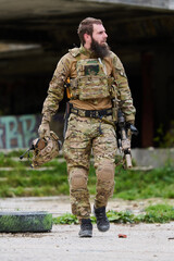 Obraz premium Bearded Soldier in Camouflage Tactical Gear Walking With Rifle and Helmet Near Abandoned Building