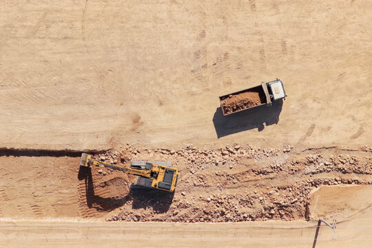 Heavy machinery including an excavator digging a trench, loading a dump truck with earth and rocks during extensive earthmoving operations at a large commercial construction site