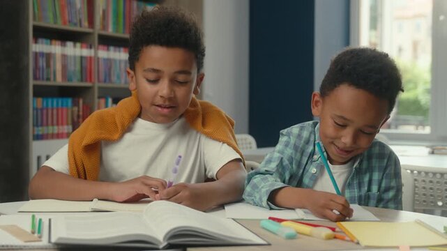 Two clever concentrated smiling little African American boys kids writing in school library at table older brother peeking correct answer looking in copybook of younger boy funny children pupils write