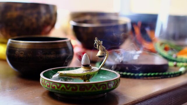 Close up of a smoking incense cone in a brass burner shaped like a small lamp, with blurred singing bowls behind. Meditation rituals and calming aromas help create quiet spaces for rest and focus.