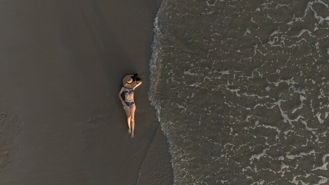 Drone view of a woman in a black bikini relaxing happily on a sandy beach.