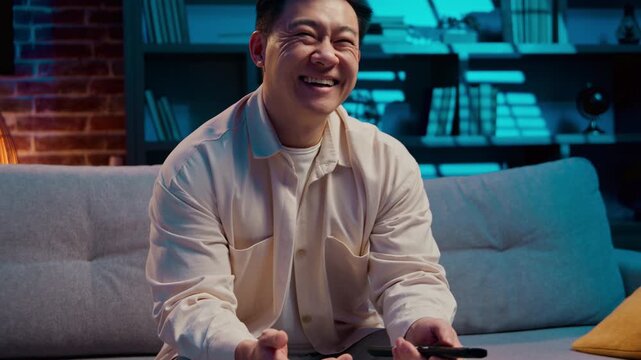 Joyful Asian middle-aged man switching channels tv laughing finding humorous program laugh at night home positive Korean Japanese Chinese male watching television funny ridiculous movie at evening