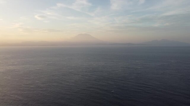 Aerial drone hovering at sunset over the ocean at Nusa Penida with distant view toward mainland Bali and volcanic mountains on the horizon, Indonesia.