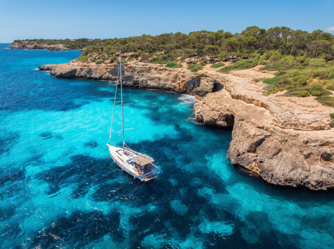 Aerial drone view of sailboat on blue sea, beautiful island, mountain, green forest on summer sunny day. Rocky sea coast, turquoise water in Sardinia, Italy. Top down view of yacht. Summer vacation