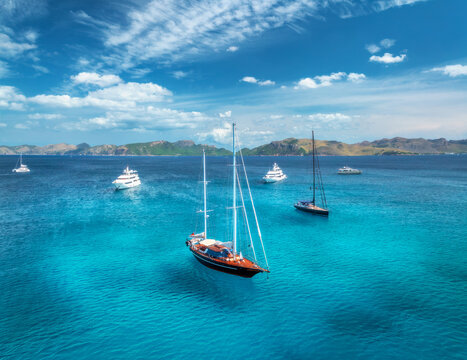Aerial drone view of yachts and sailboats anchored in crystal clear turquoise sea near coast and mountains, sky with clouds. Mallorca, Balearic Islands, Spain. Mediterranean summer travel. Top view